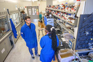Two undergraduate researchers wearing blue lab coats stand facing each other in a lab
