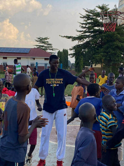 A man in a blue Notre Dame Football 2022 t-shirt and white pants with a green whistle gestures to a group of diverse children on an outdoor basketball court. A building with solar panels and a basketball hoop with a red net are visible in the background under a cloudy sky.