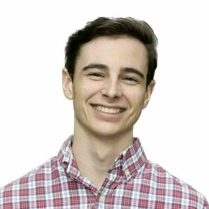 A young man with dark hair smiles broadly, showing teeth, while wearing a red, white, and blue plaid button-down shirt against a plain white background.