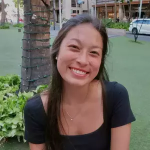 A young woman with long dark hair, freckles, and pearl earrings smiles widely at the camera, showing teeth. She wears a black top and a silver necklace with a small pendant. Behind her are a palm tree wrapped in string lights and green grass.