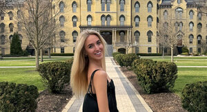 Young woman with blonde hair smiles over her shoulder on a path towards Notre Dame's iconic Main Building.