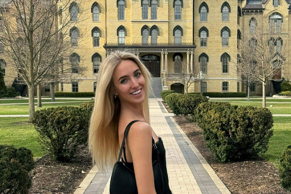 Young woman with blonde hair smiles over her shoulder on a path towards Notre Dame's iconic Main Building.