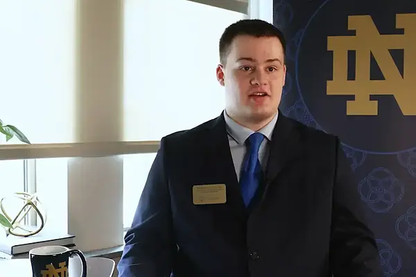Benjamin Gallei in a dark suit and blue tie speaks, with a gold Notre Dame logo on a blue banner behind him.