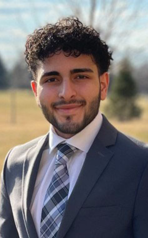 A young man with dark, curly hair and beard, wearing a grey suit, white shirt, and plaid tie, smiles gently outdoors.