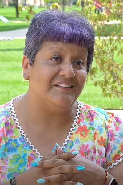 Esther Chavez is a woman with short purple hair and a colorful floral top clasps her hands, smiling gently in a sunny outdoor setting.