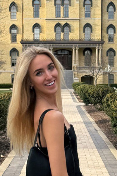 Smiling blonde woman in a black top on a campus path, with Notre Dame's yellow Main Building in the background.