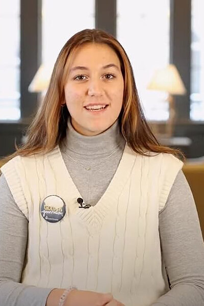 Smiling woman with brown hair and highlights, wearing a grey turtleneck and cream vest with a "Golden Generation" pin in a campus lounge.