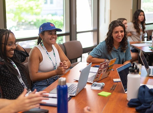 Notre Dame Pre-college students laugh and engage around a wooden table with laptops.
